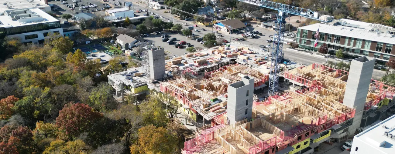 Aerial view of a construction site with a large crane and partially built structures surrounded by trees. Nearby roads and a suburban area in the background.