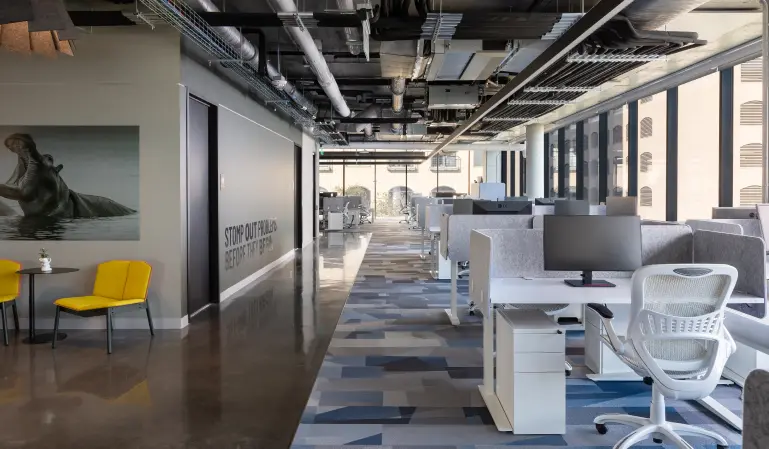 Modern office interior with open-plan workstations, grey dividers, and ergonomic chairs. Bright space with large windows, yellow chairs, and artwork.