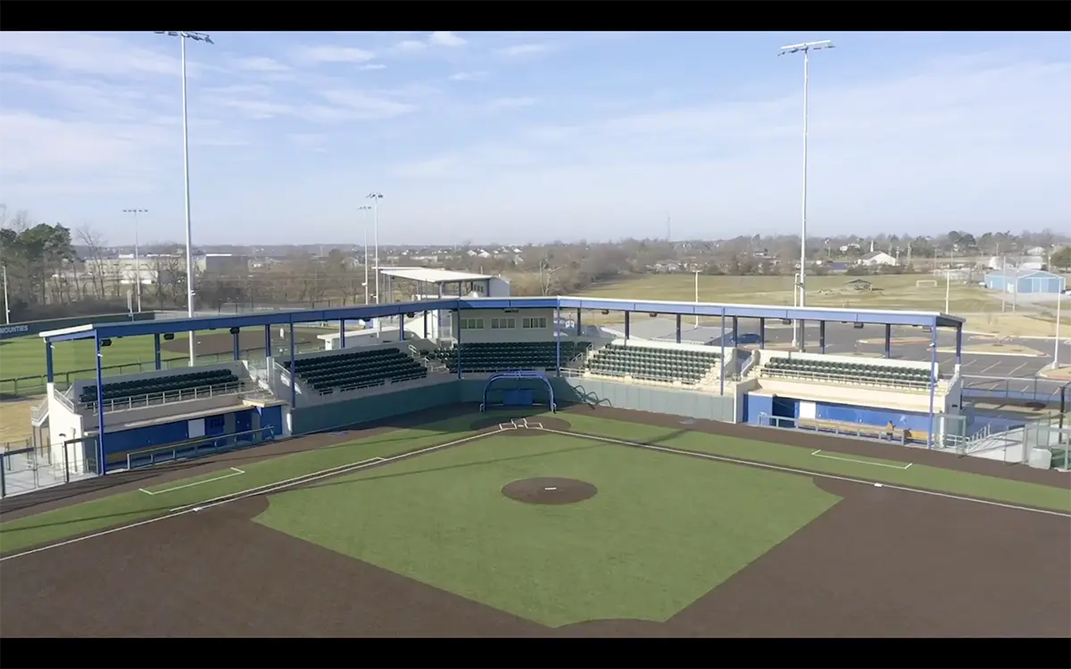 Aerial view of an empty baseball field with green and brown turf, surrounded by blue seating stands. The sky is clear, suggesting a peaceful atmosphere.