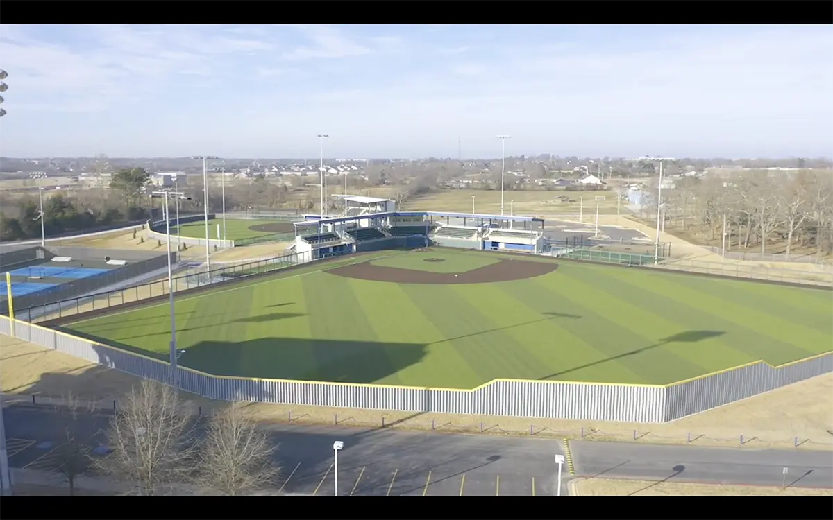 Aerial view of a well-maintained, empty baseball field with striped grass and a dirt infield. Bleachers and a few trees are visible in the distance under a clear sky.