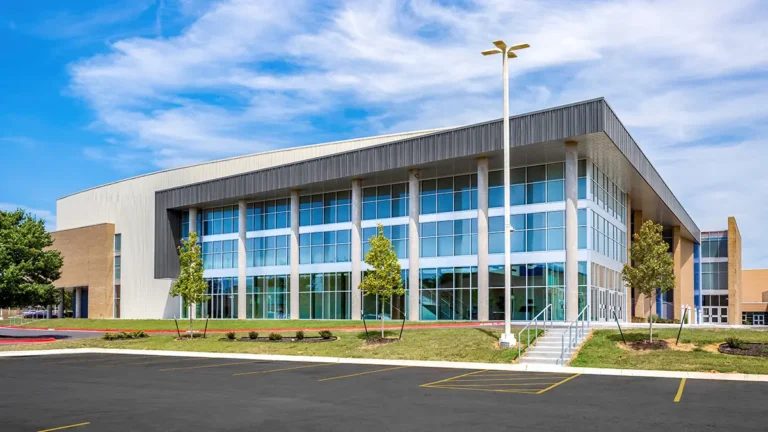 Modern two-story building with large glass windows, surrounded by small trees and a well-kept lawn, under a clear blue sky. The atmosphere is calm and orderly.
