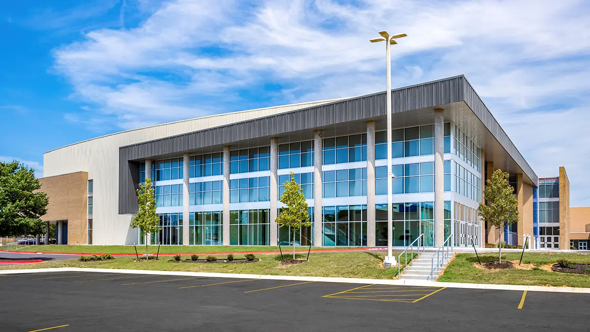 Modern two-story building with large glass windows, surrounded by small trees and a well-kept lawn, under a clear blue sky. The atmosphere is calm and orderly.
