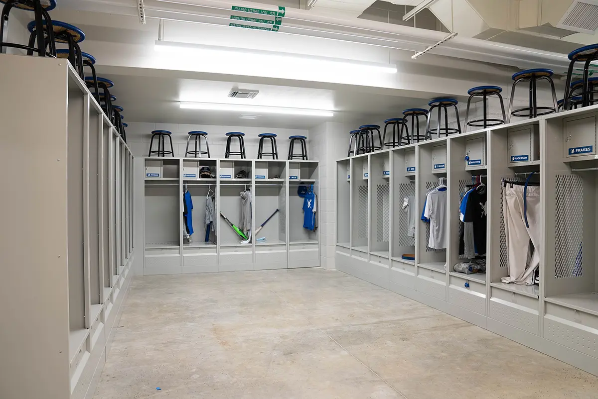 Spacious locker room with light gray lockers lining the walls, blue and white sportswear, equipment, and stools on top, under bright fluorescent lights.