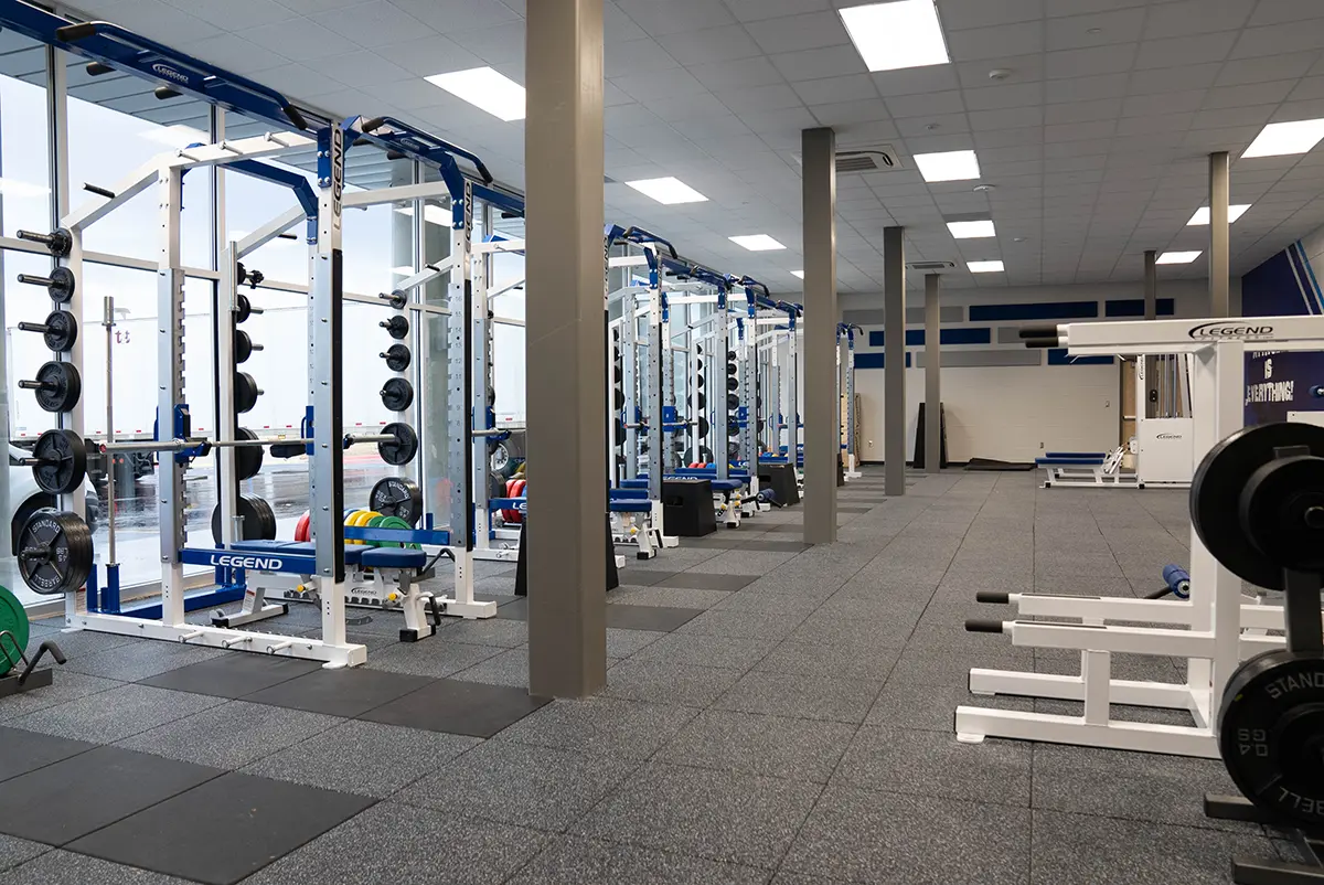 A spacious gym with weightlifting racks lined up against glass walls. The gym is well-lit, featuring various weights and equipment, creating an organized and motivational atmosphere.