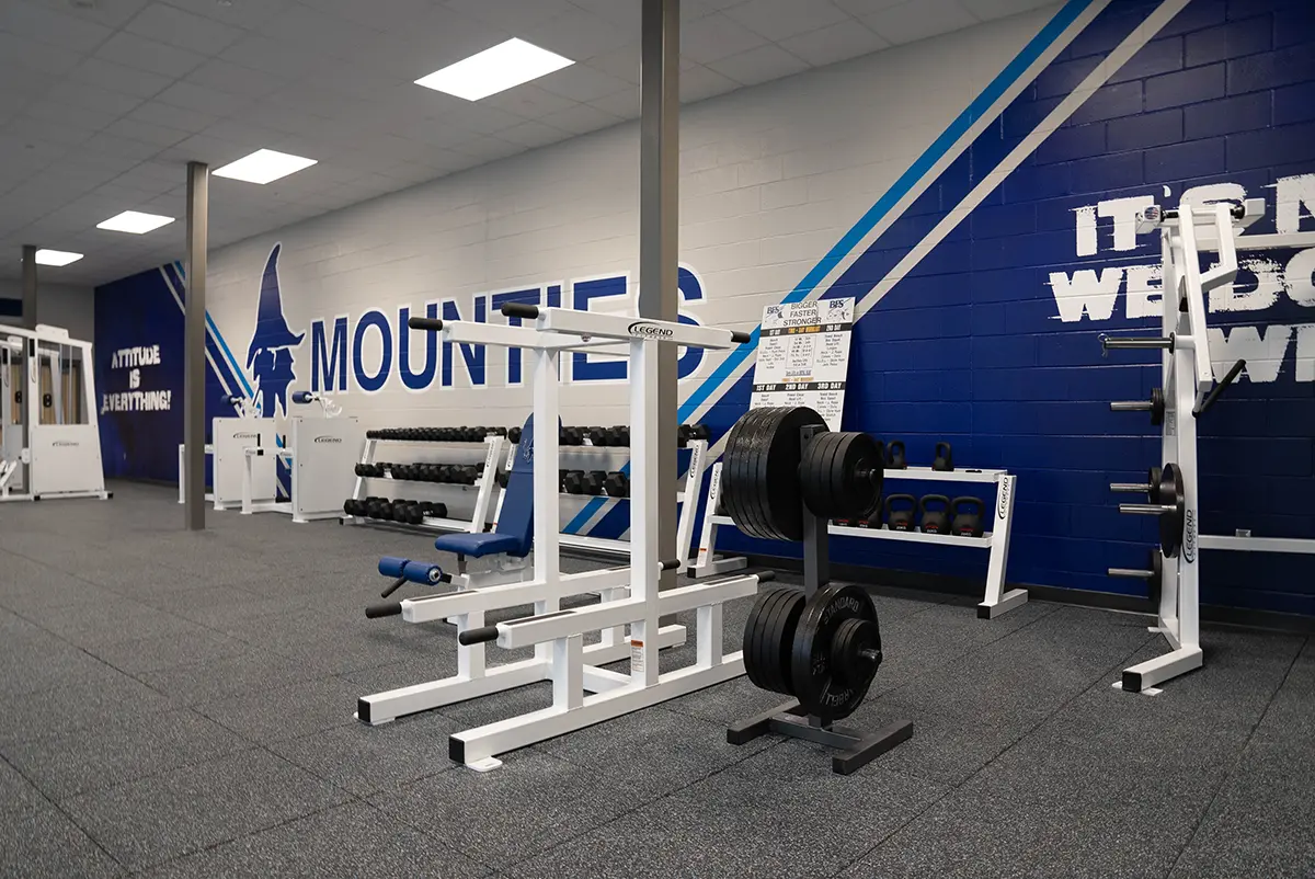 Modern gym interior with weightlifting equipment and dumbbells. The walls feature motivational quotes and the word “Mounties” in bold letters. The space feels energetic and motivating.