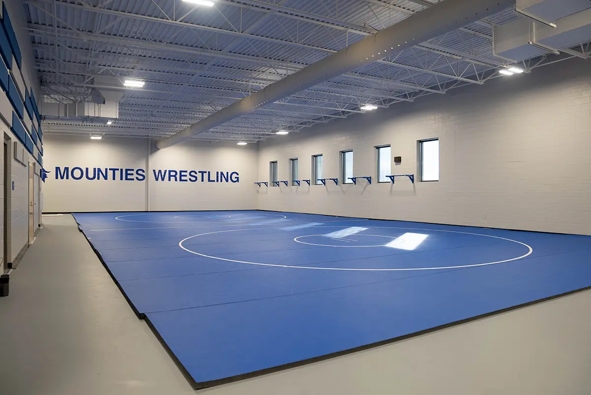 A spacious, modern wrestling gym with blue mats covering the floor. "Mounties Wrestling" is painted on the wall, with windows to the right. Bright lighting.
