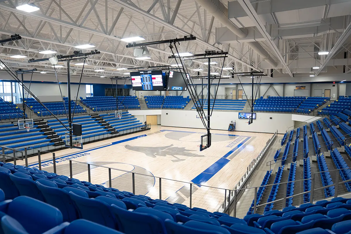 Empty indoor basketball court with polished wood flooring and blue seating. Overhead, scoreboard displays. Bright lighting and modern design create a spacious feel.