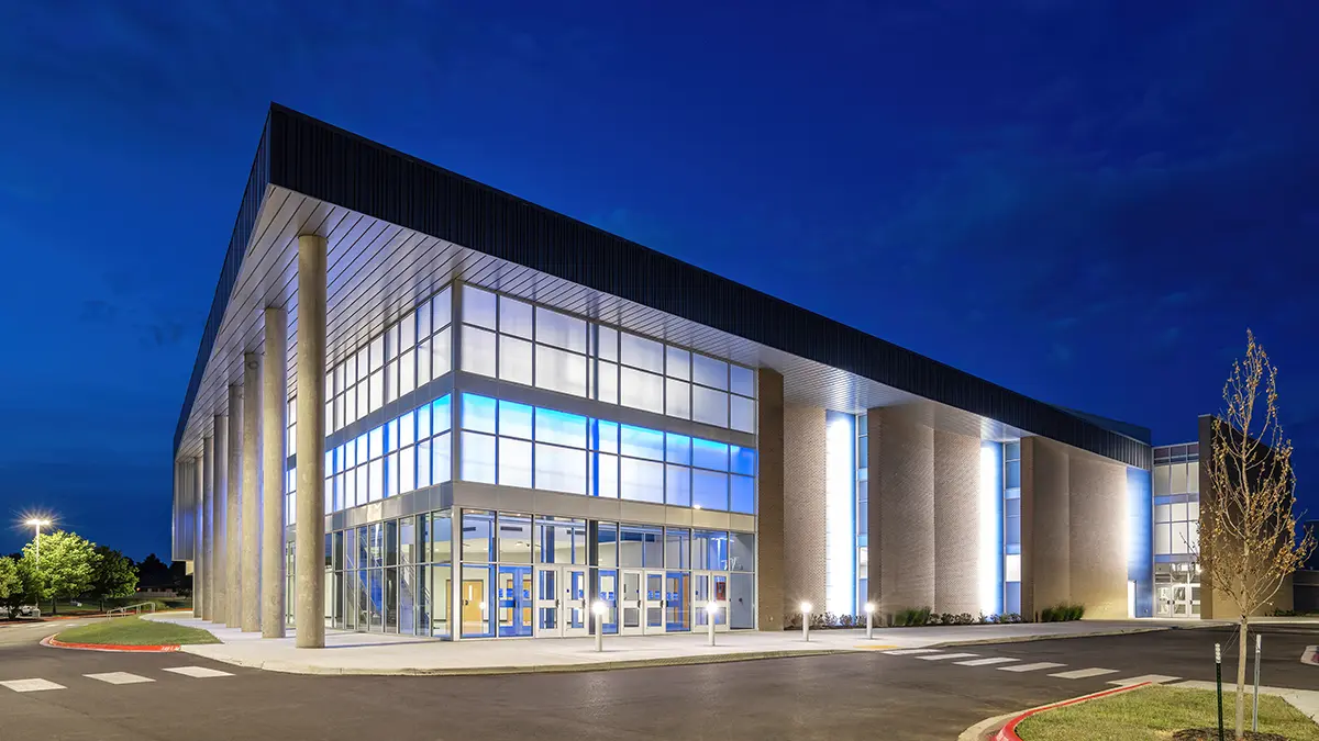 Modern building at night with large glass windows emitting a blue glow. The structure features tall columns and is surrounded by a quiet parking area.