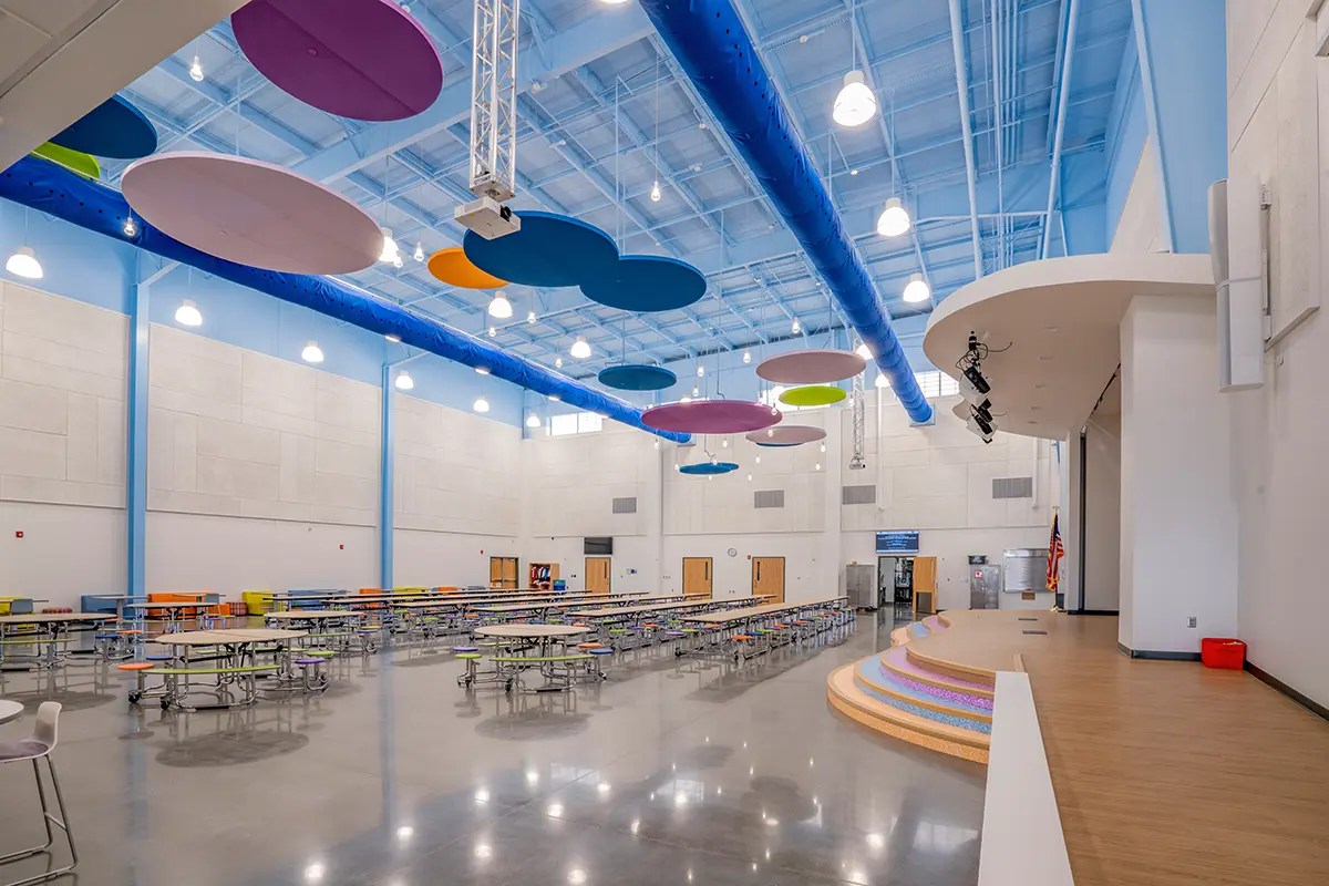 Spacious school cafeteria with colorful circular ceiling panels and bright blue beams. Rows of tables fill the room, and a stage with rainbow steps is on the right.