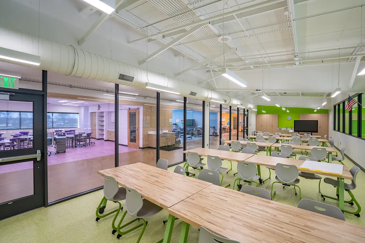 Modern classroom with large wooden tables, gray chairs, and green accents. Glass walls reveal adjacent rooms. Bright, spacious, and orderly.