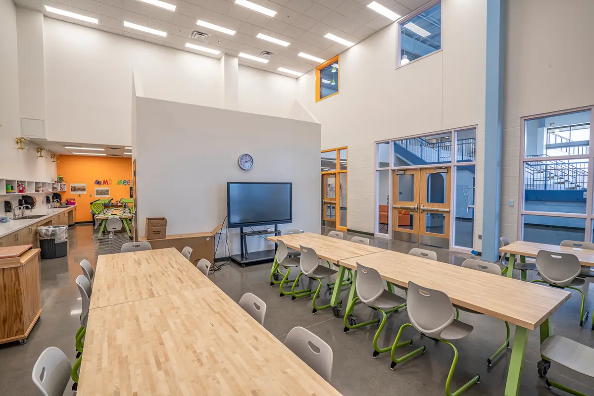 Spacious classroom with light wooden tables and gray chairs. A digital screen and clock are on a wall. Bright orange accents add a lively touch.