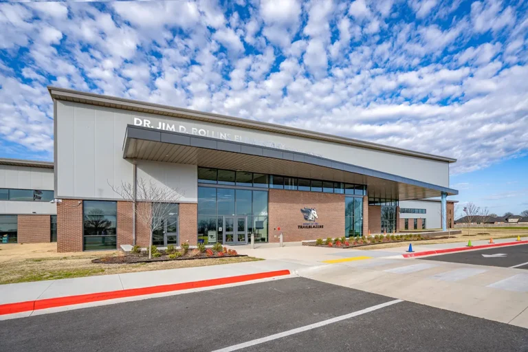 Modern school building with large glass windows and a brick facade under a vibrant, cloudy sky. The entrance is wide and welcoming.