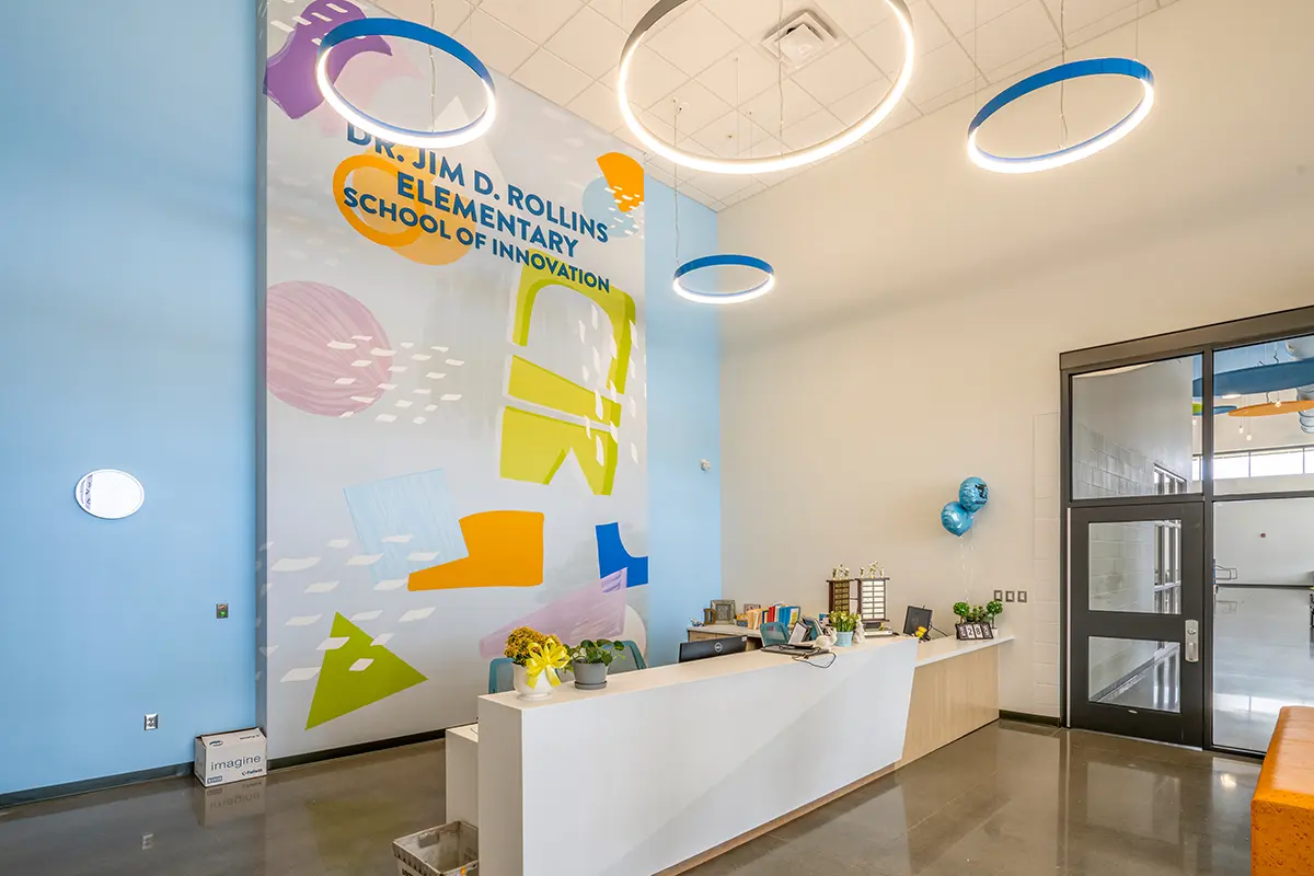 Bright school lobby with a modern design; features a reception desk, vibrant wall art with geometric shapes, flowers, and large circular lights overhead.