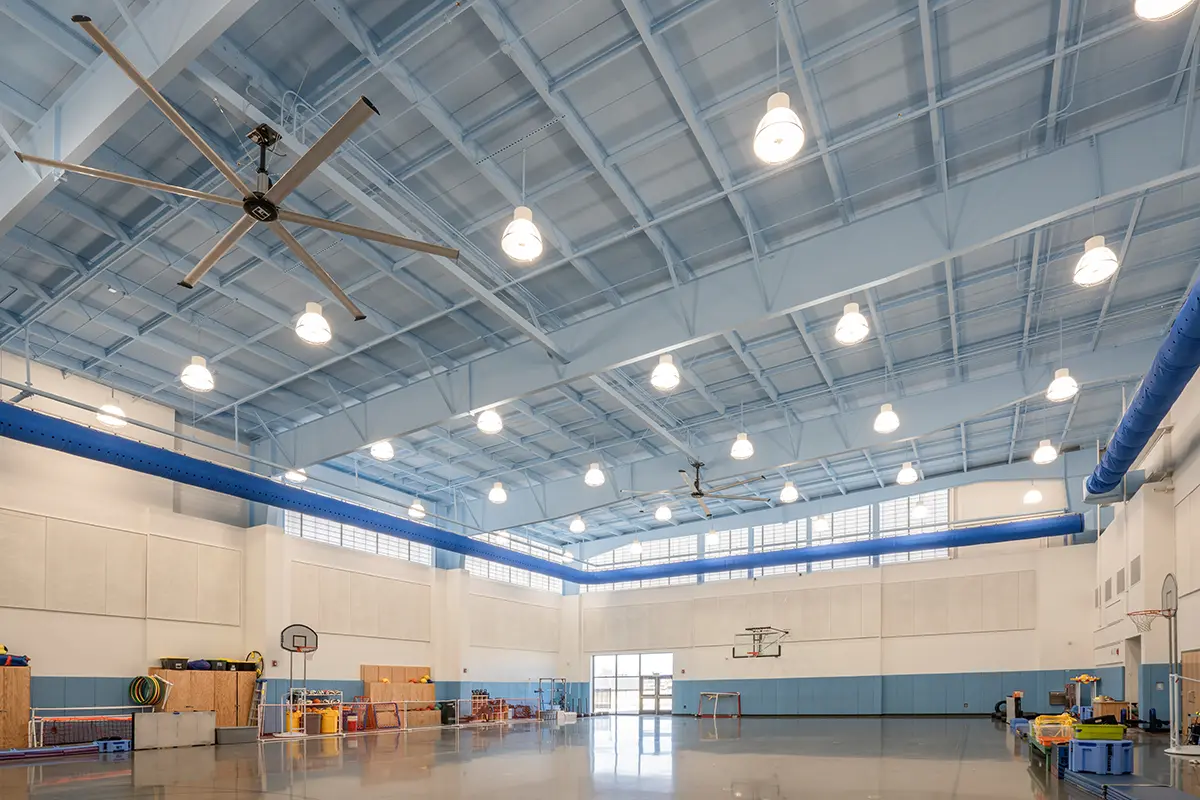 Spacious gymnasium with high ceiling and large lights. Basketball hoops, colorful gym equipment, and a fan are visible. Bright and organized atmosphere.