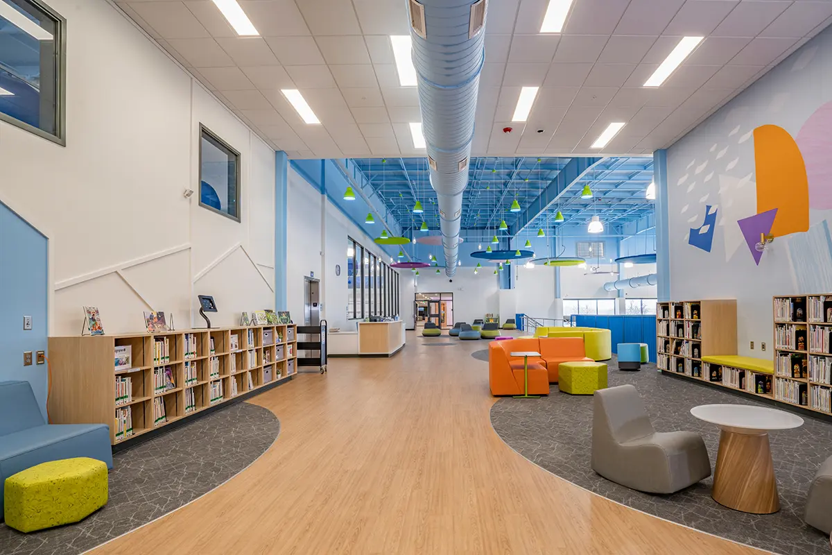 A spacious, colorful library with modern decor. Bookshelves line the walls, vibrant seating options, and geometric ceiling art create an inviting, cheerful atmosphere.