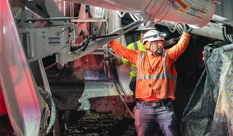 A construction worker in an orange safety vest and helmet operates machinery at night, conveying focus and determination in a dimly lit environment.