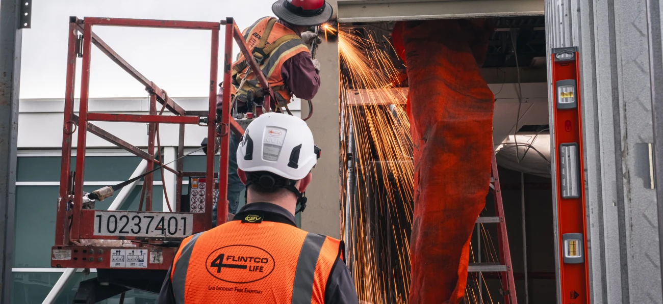 A construction worker uses a grinder on a building facade, creating bright sparks. Another worker observes, wearing safety gear and an orange vest.