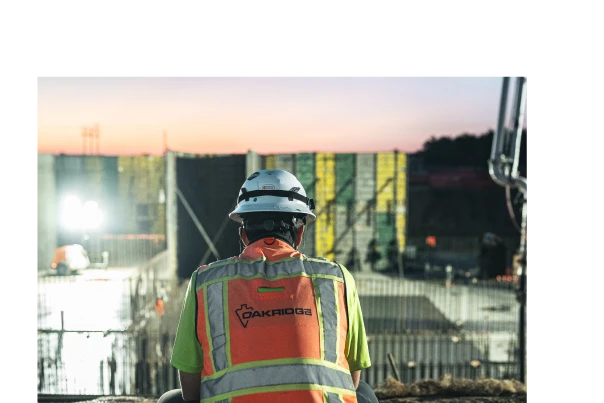 Construction worker in a helmet and safety vest overlooks a construction site at dusk. The site is illuminated with artificial lights, creating a focused, industrious atmosphere.