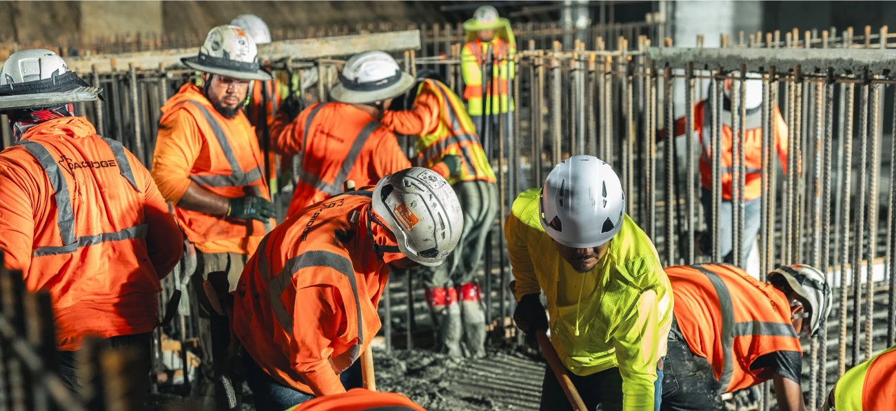 Construction workers in bright orange safety vests and helmets pour and smooth concrete in a rebar-framed area, showing teamwork and focus in a busy worksite.