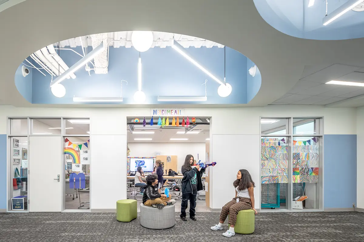 Modern classroom with colorful decor, featuring three students interacting. Bright, open space with rainbow flags and abstract art. Energetic, welcoming atmosphere.