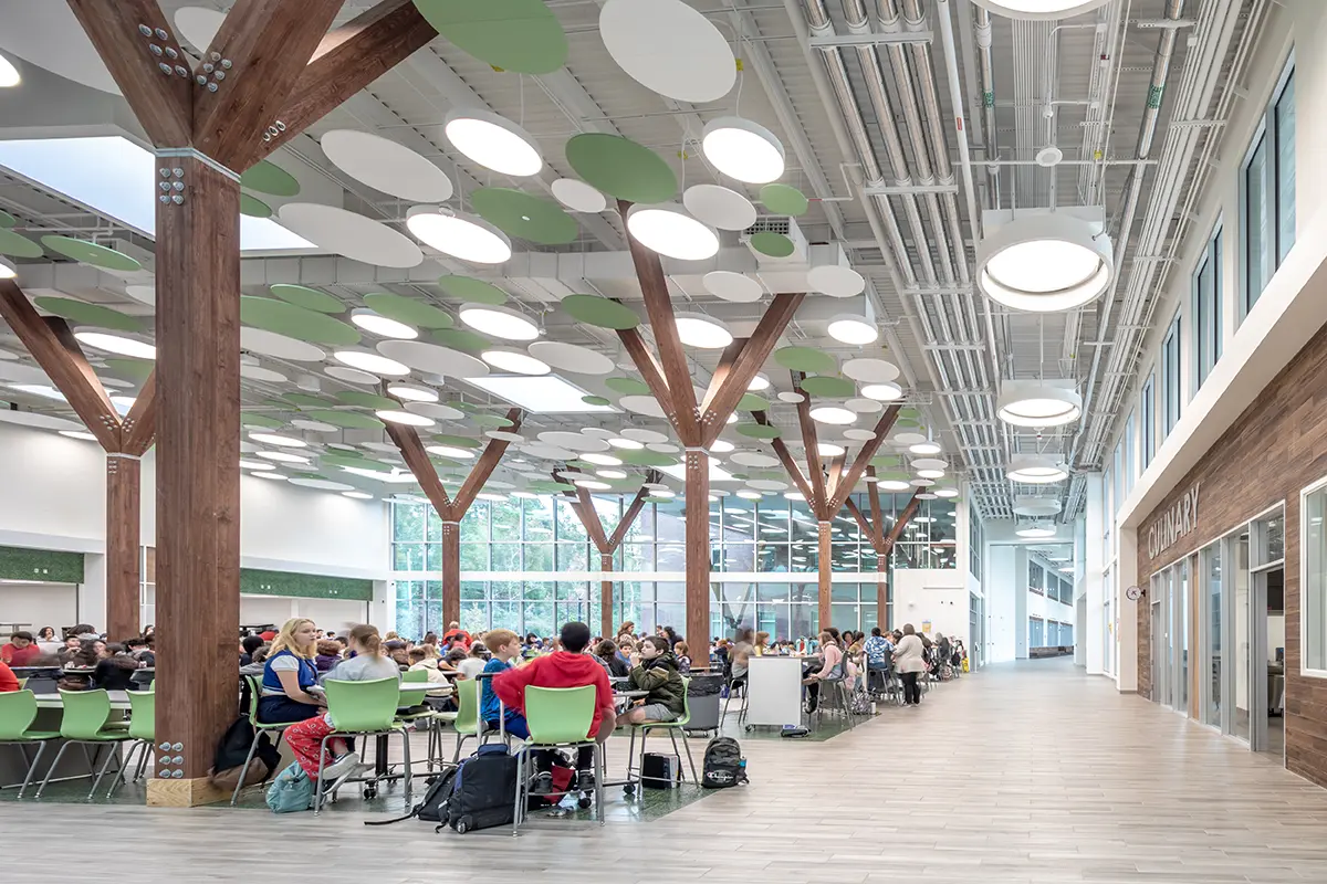Modern cafeteria with high ceilings and tree-like wooden columns. Circular green and white designs hang from the ceiling. People sit at tables, creating a lively atmosphere.