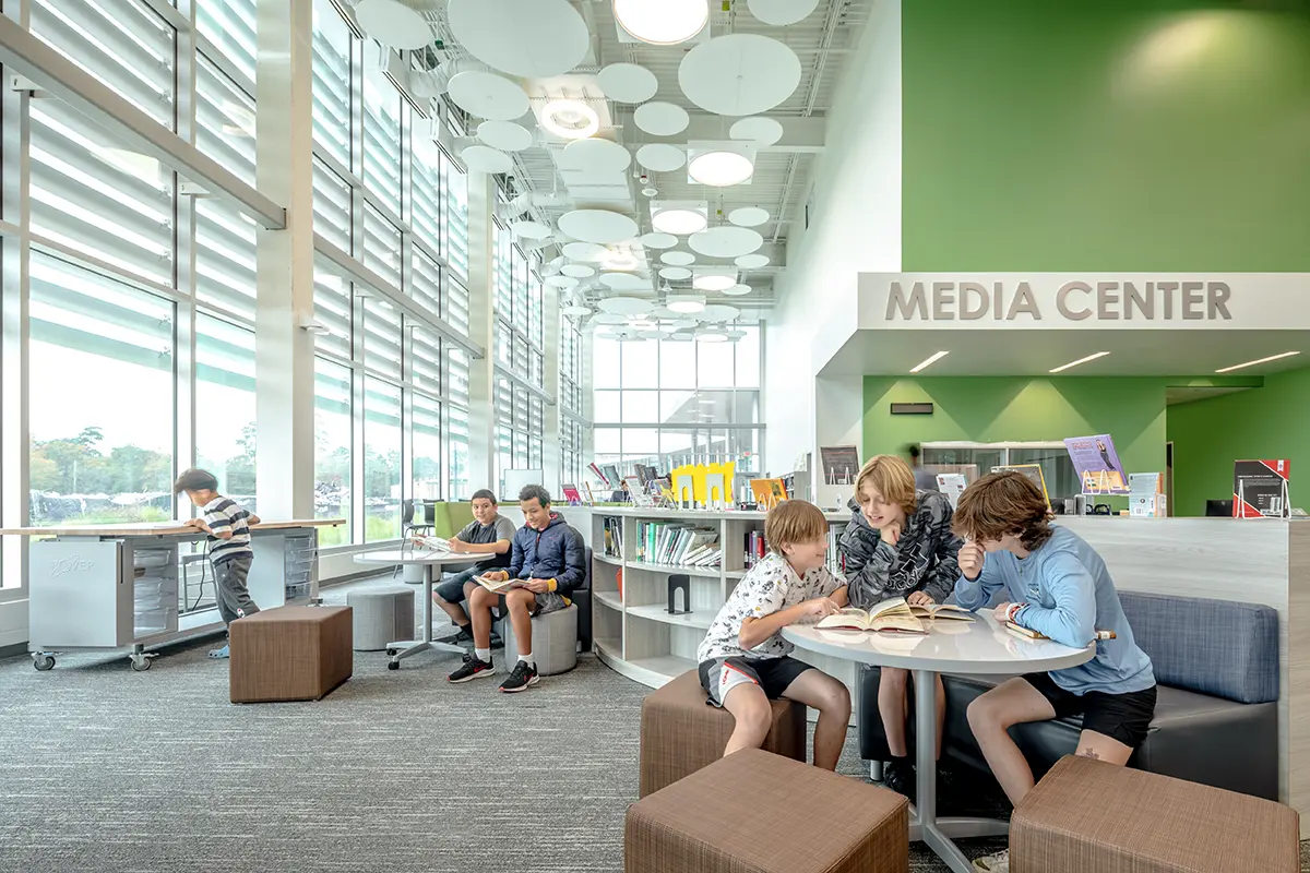 Modern media center with large windows, green walls, and circular ceiling lights. Five kids seated on stools read books, creating a focused atmosphere.