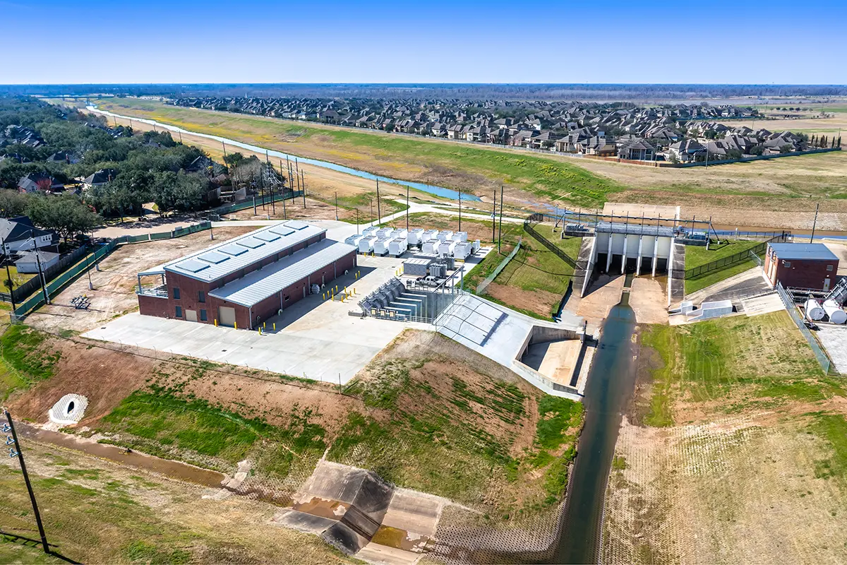 Aerial view of an industrial facility with red brick buildings and machinery, adjacent to a drainage canal. Surrounded by fields and a residential area.