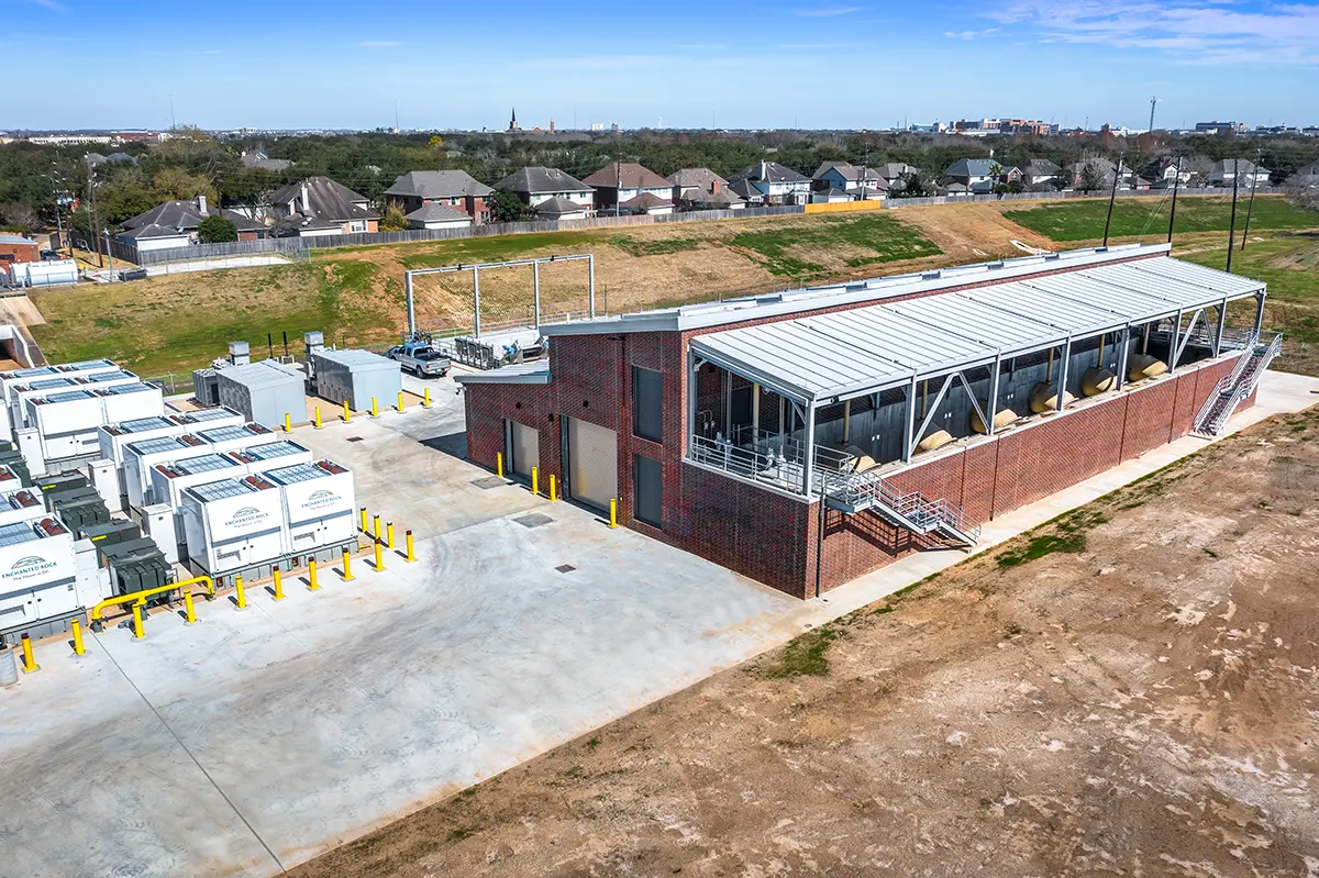 Aerial view of a large industrial facility with a brick building and modular units on a concrete lot. Nearby, a residential area and open land are visible.