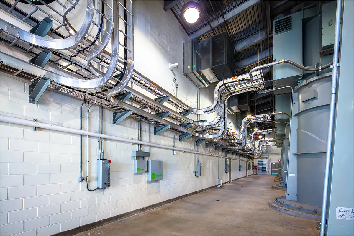 A brightly lit industrial corridor with numerous metal cables and conduits running along the ceiling and walls, conveying technological complexity.
