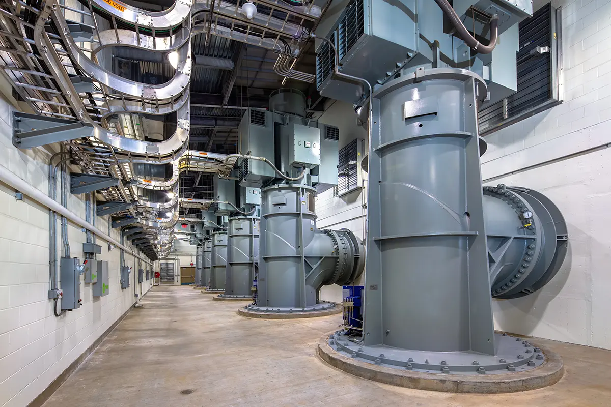 A spacious industrial room with large, cylindrical gray turbines lined up, overhead metal pipes, and concrete flooring, conveying a high-tech, functional tone.