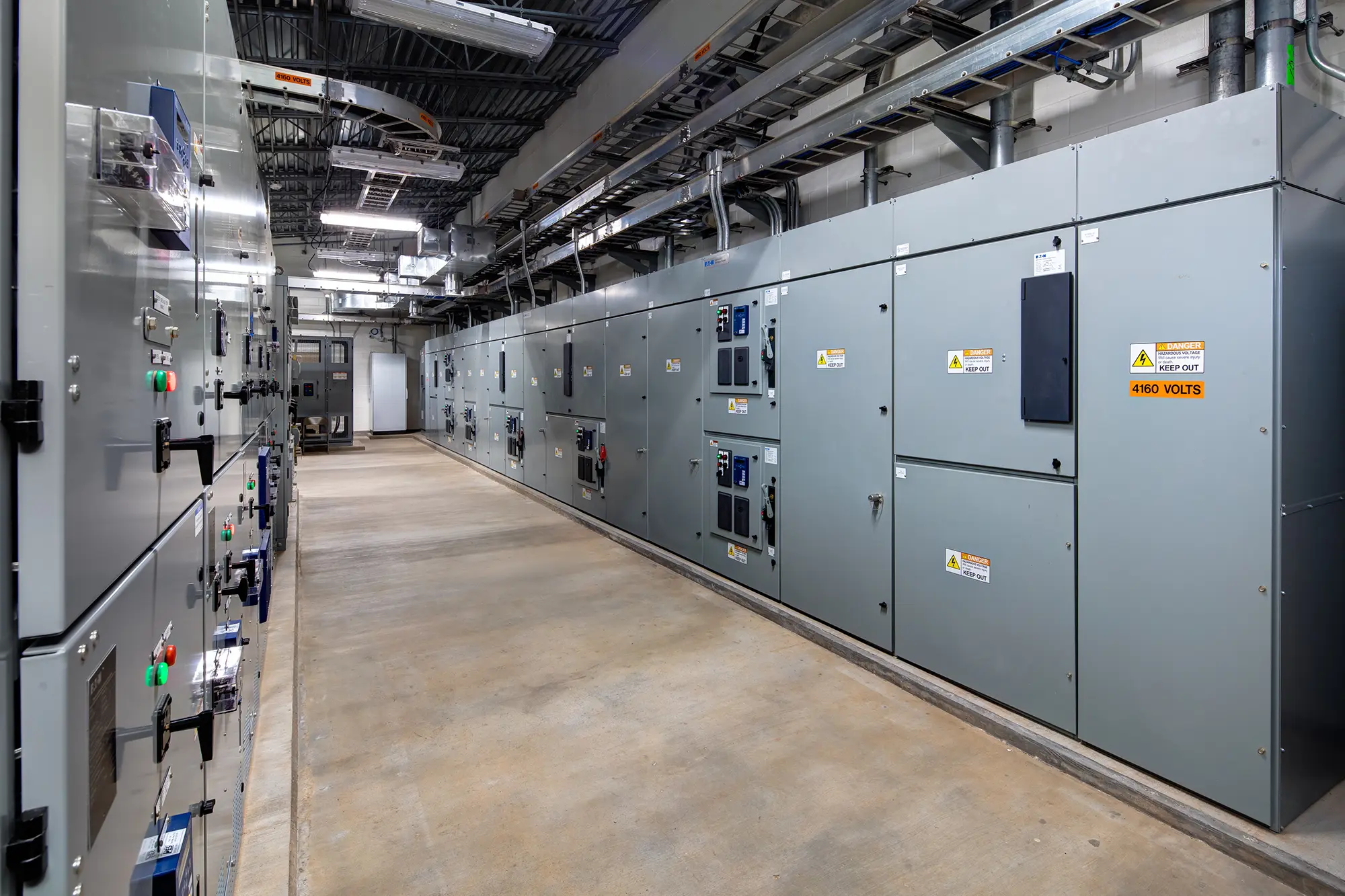 A corridor in an industrial facility is lined with large gray electrical panels on both sides. Warning signs read "480 VOLTS." The setting is clean and organized.