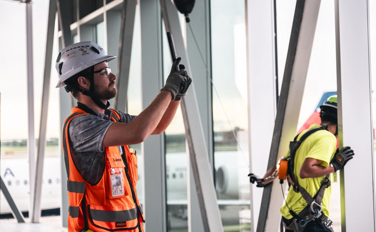 Aerial view of a construction site with a large crane and partially built structures surrounded by trees. Nearby roads and a suburban area in the background.