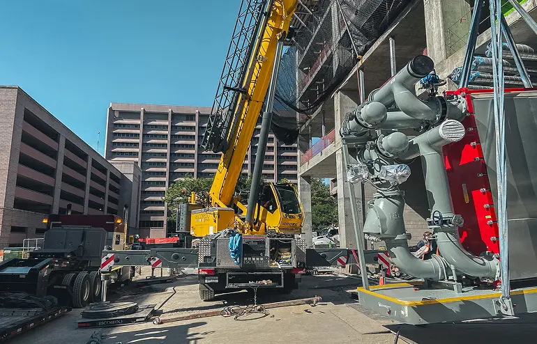 Yellow crane lifting large mechanical equipment next to a partially constructed building. Nearby, a multi-story parking garage and trees are visible under a clear blue sky.