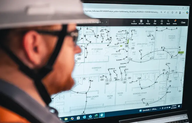 A person wearing a white hard hat and glasses observes a detailed technical blueprint on a computer screen, conveying focus and precision in a professional setting.