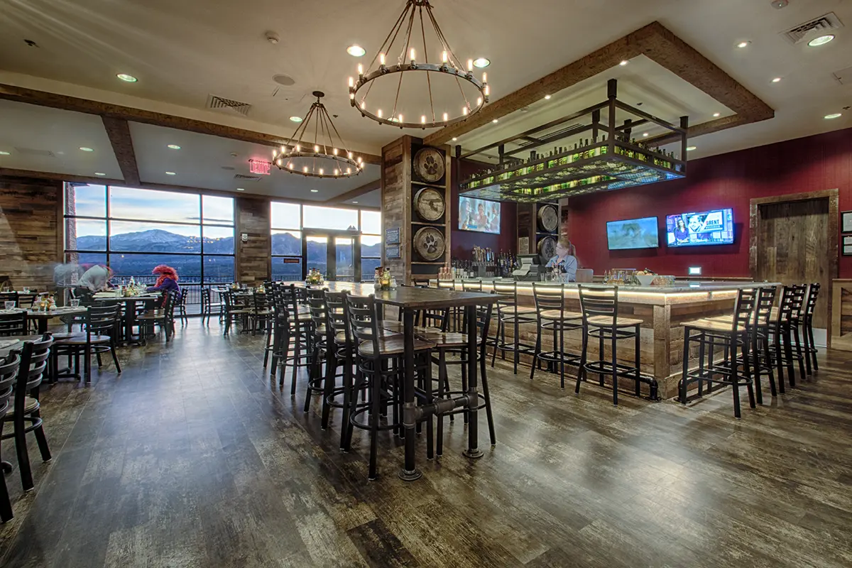 Rustic restaurant interior with wooden furniture, chandeliers, and a central bar. Large windows showcase mountain views. Cozy and inviting atmosphere.