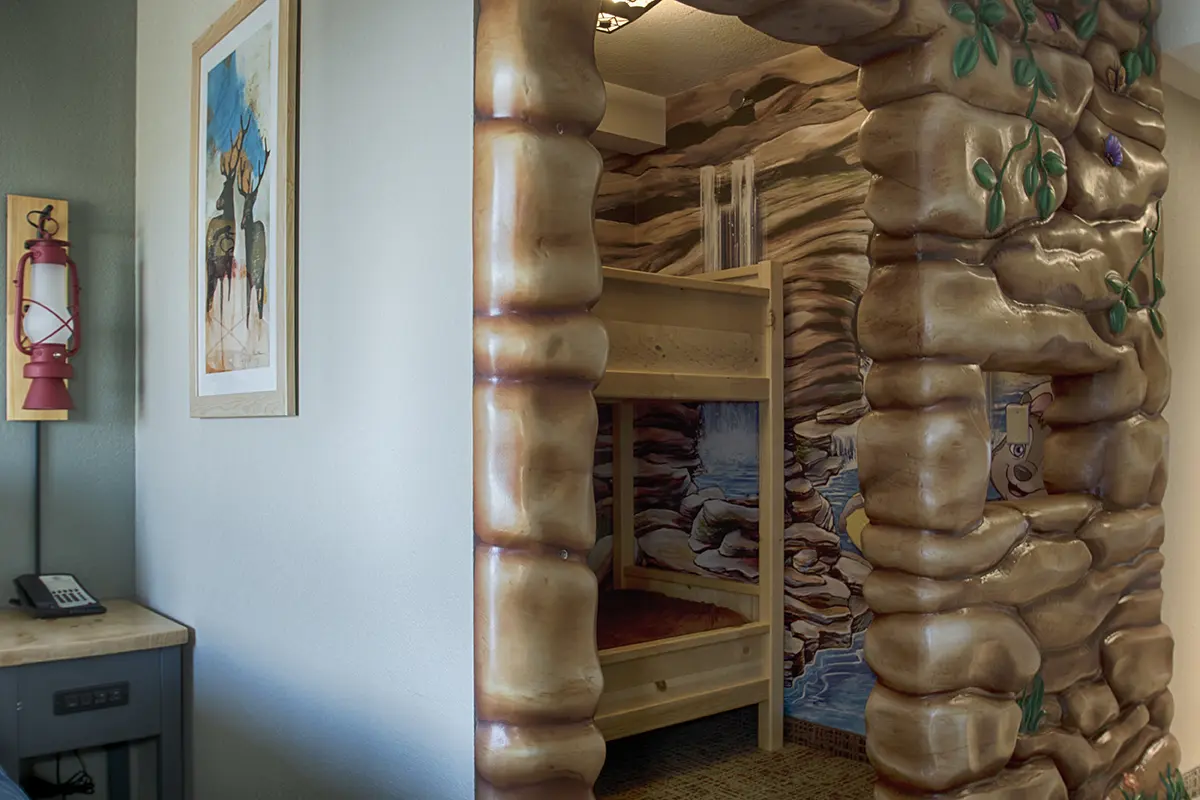 Hotel room with a playful cave-themed bunk bed area. The entrance is styled as rocks with leaves, next to a painting and a red wall lantern. Cozy and imaginative.