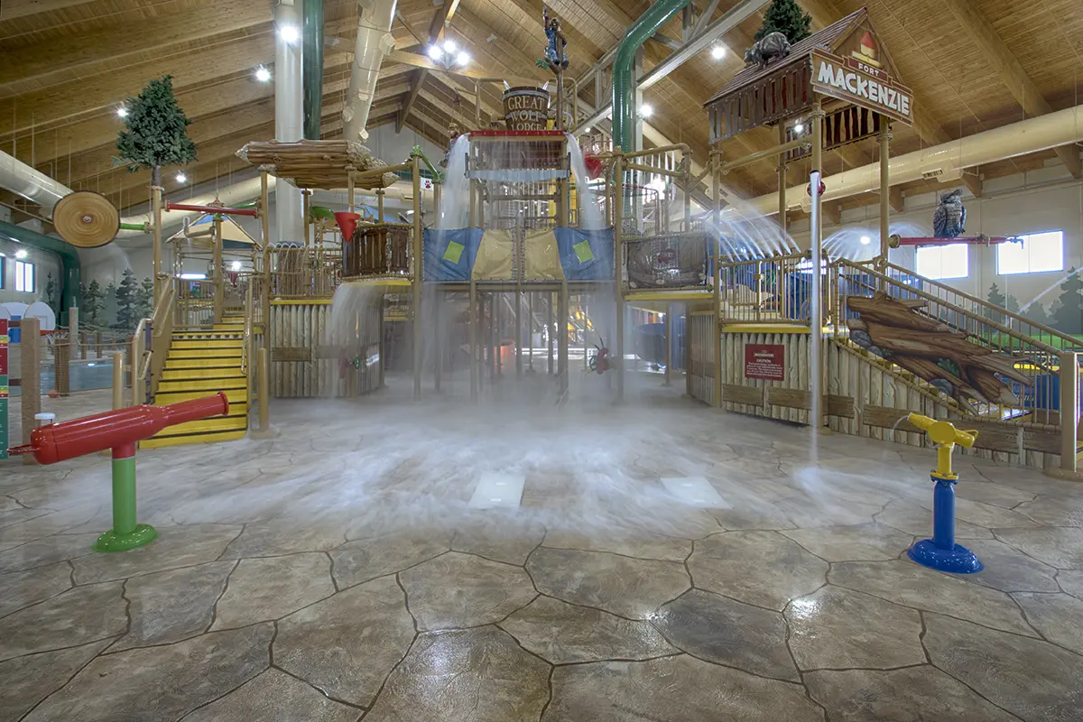 Indoor water park with wooden-themed play structure, water cannons, slides, and overhead buckets spilling water. Bright, playful atmosphere.