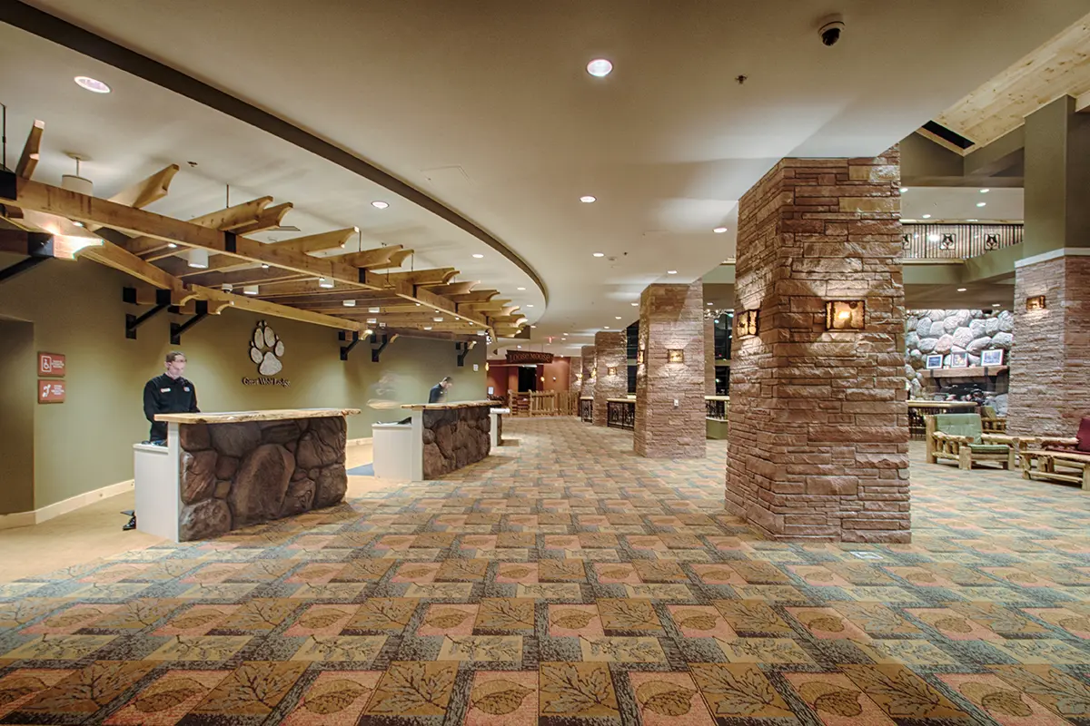 Spacious hotel lobby with wooden and stone accents, colorful patterned carpet, curved check-in desks, and warm lighting, creating a welcoming atmosphere.