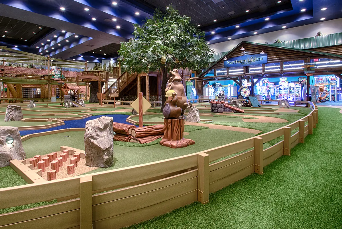 Indoor mini-golf course with bear sculpture, faux trees, and rustic decor. Arcade in the background, bright lighting, playful and inviting atmosphere.