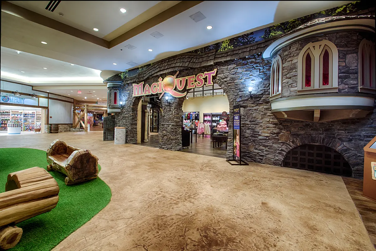 Fantasy-themed entrance to “MagiQuest” with stone castle facade and archway. Indoor setting with cozy log benches on green turf nearby. Inviting and adventurous.