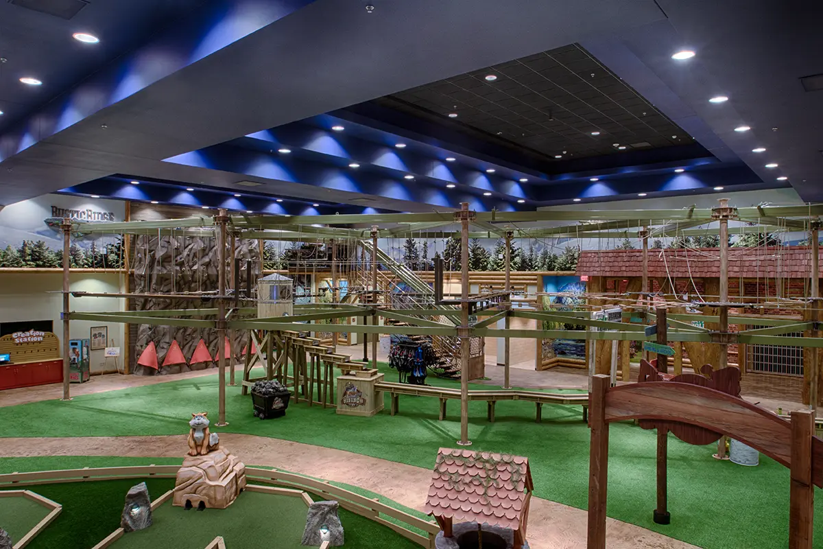An indoor adventure park with rope bridges and climbing structures. Green turf covers the floor, while colorful artificial trees and play areas create a playful atmosphere.