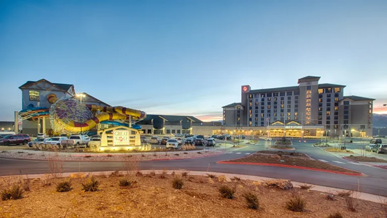 A resort with a tall hotel and vibrant water slides at dusk. The foreground shows a lit entrance sign and parked cars under a clear evening sky.