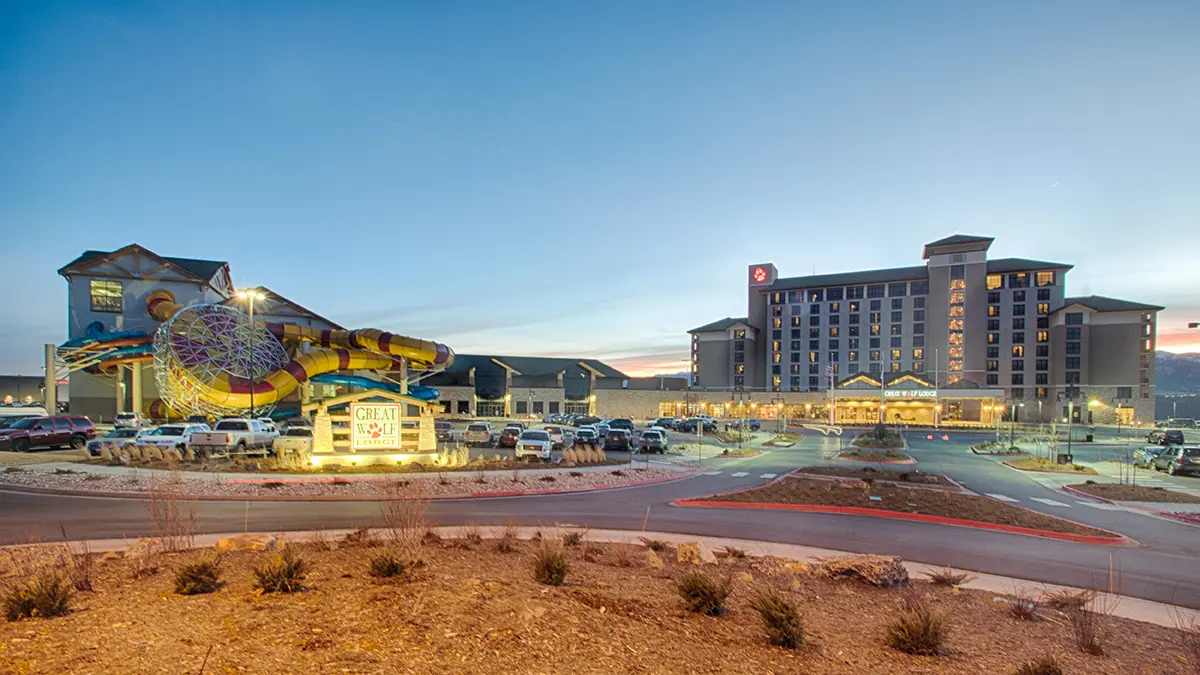 A resort with a tall hotel and vibrant water slides at dusk. The foreground shows a lit entrance sign and parked cars under a clear evening sky.