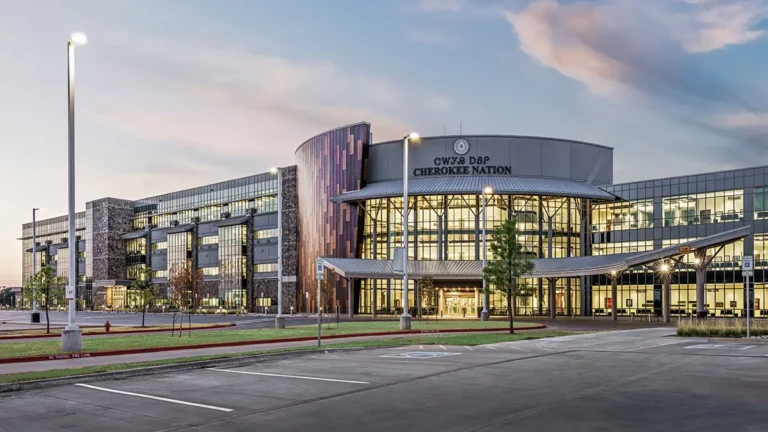 Modern building at sunset featuring glass and stone facade, curved roof with bold text, surrounded by a calm parking area and soft blue-pink sky.