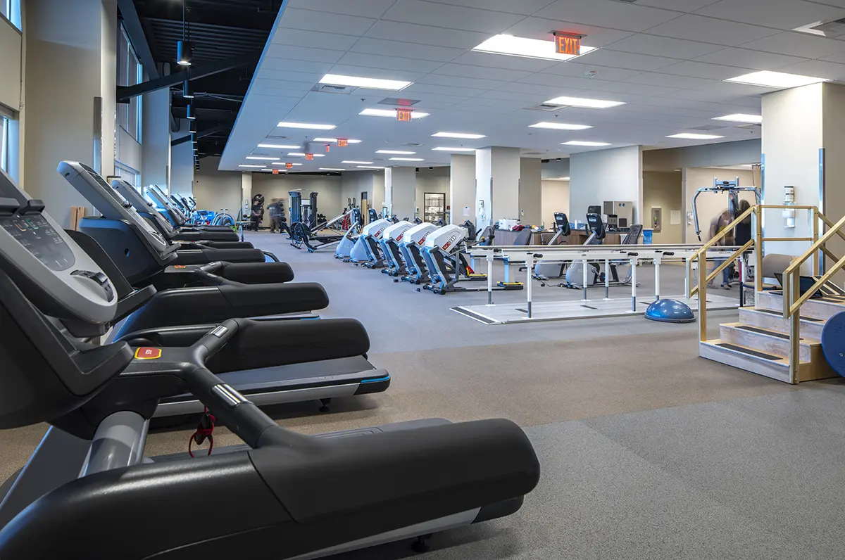 A spacious, well-lit gym with rows of treadmills and exercise bikes on a carpeted floor. Physical therapy equipment and steps are visible. Calm, professional atmosphere.