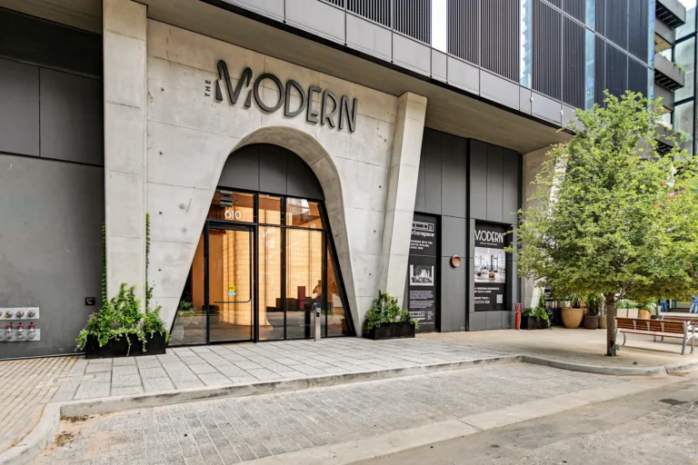Modern building entrance featuring sleek glass doors under an arched concrete facade. Potted plants and a tree add greenery, creating an inviting atmosphere.