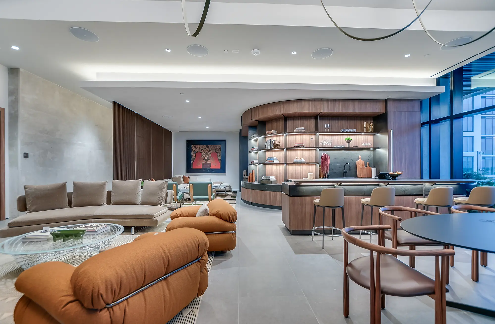 세련된 가구, 곡선형 소파, 갈색 라운지 체어, 바 공간이 조화를 이루는 현대적인 생활 공간. 벽면에는 선반이 장식되어 있으며, 커다란 창문으로 자연 채광이 들어온다.