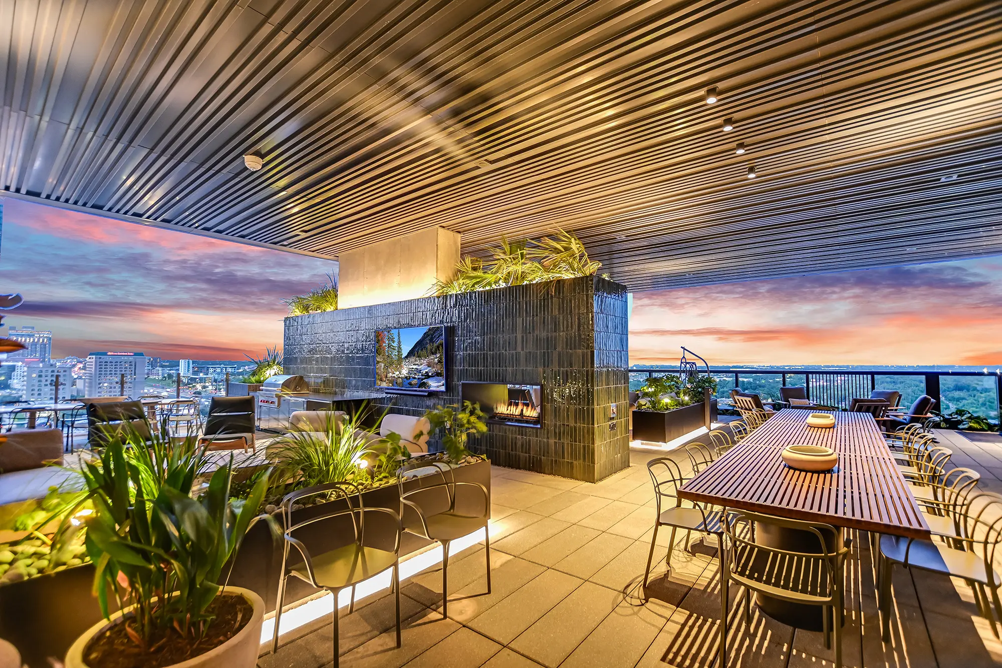 일몰을 배경으로 한 고급스러운 루프탑 테라스에는 현대적인 가구, 긴 식탁, 무성한 식물, 세련된 벽난로가 자리 잡고 있습니다. 도시의 스카이라인이 한눈에 들어옵니다.