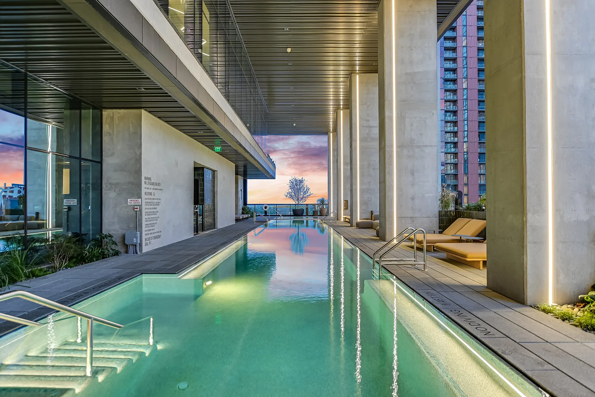 빛나는 물과 세련된 디자인의 현대식 실내 수영장. 라운지 의자와 높은 기둥이 양옆을 둘러싸고 있다. 유리 너머로 보이는 석양 풍경이 고요한 분위기를 자아낸다.