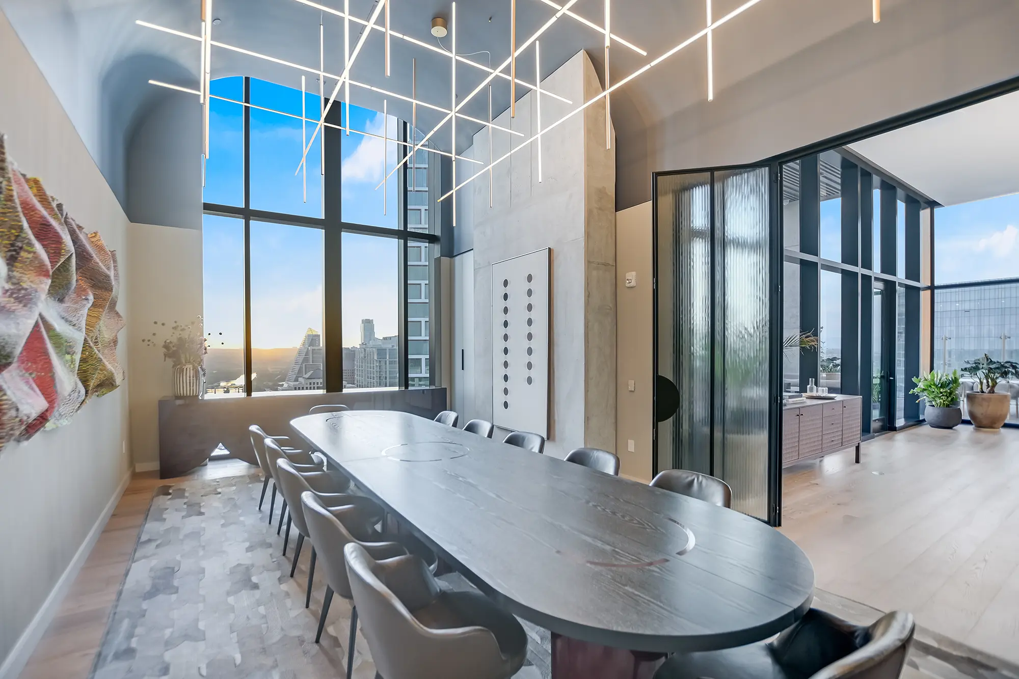 넓고 현대적인 식당에는 긴 나무 테이블과 고급스러운 의자가 놓여 있으며, 기하학적인 천장 조명이 비추고 있다. 커다란 창문 너머로 도시 풍경이 펼쳐진다.